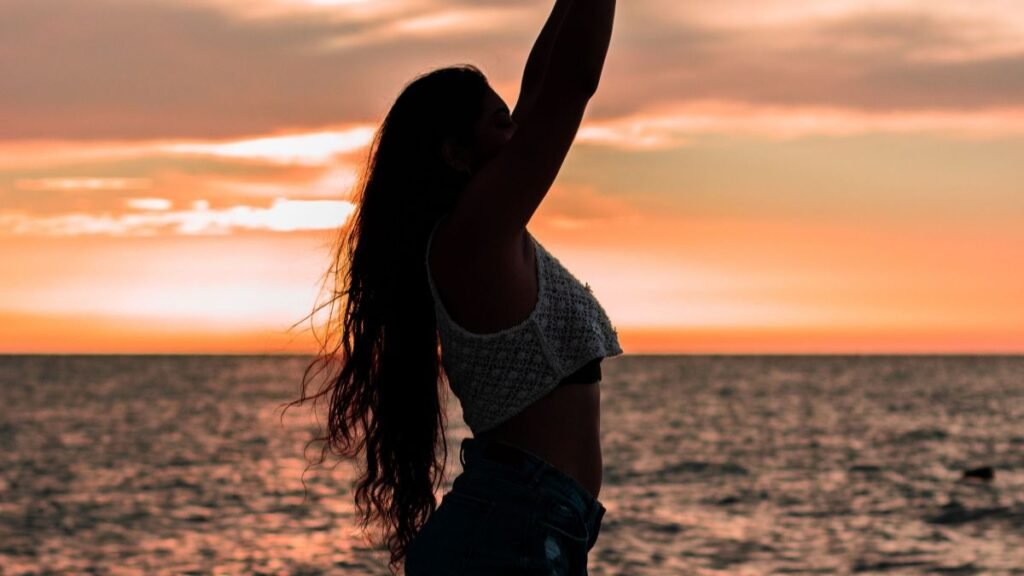 Woman standing on the beach with arms raised, embracing the golden hour
