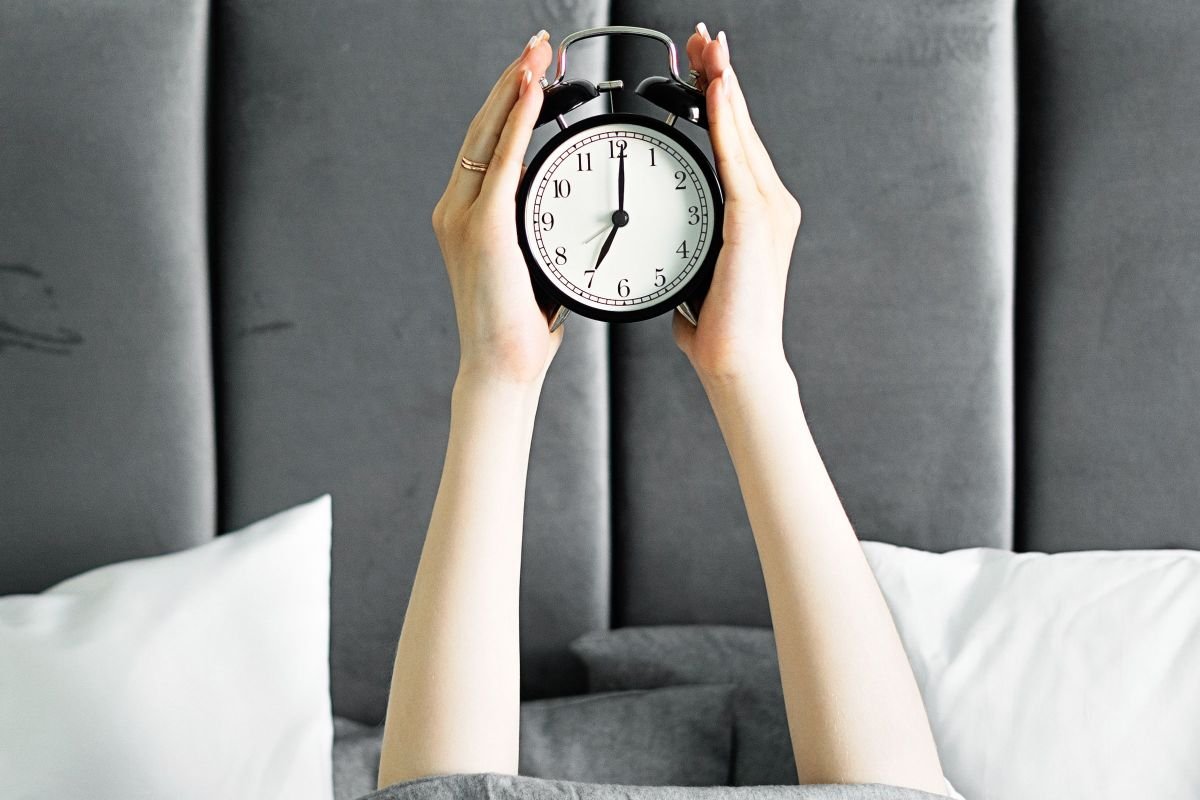 Hands holding an alarm clock above a cozy bed, representing a consistent bedtime routine